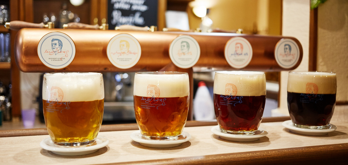 Čtyři řemeslná piva s bohatou pěnou na dřevěném baru před měděným výčepem s logy, teplá útulná hospodská atmosféra