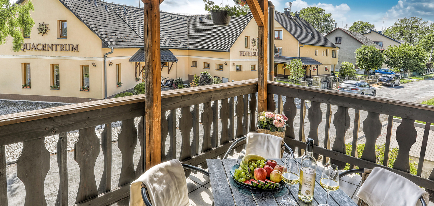 Slunný balkon rodinného apartmánu v Hotelu Slunce Rýmařov v Jeseníkách s posezením pro dva, vínem, ovocem a výhledem na hotel a Wellness & Aquacentrum Slunce.