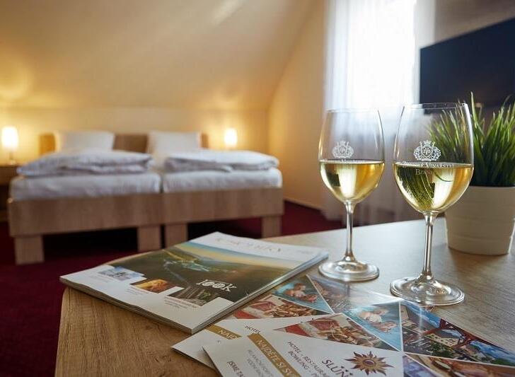 Útulný podkrovní pokoj se dvěma lůžky a tlumenými nočními lampami; na stole dvě sklenky bílého vína, brožury a květina v květináči