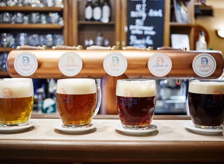 Čtyři čerstvě čepovaná piva s bohatou pěnou na dřevěném baru v restauraci pivovaru Morous v Hotelu Slunce v Rýmařově v Jeseníkách. Ochutnávka řemeslných piv z rodinného pivovaru Morous, měděný výčepní kohout a tabule s nabídkou piv v pozadí.