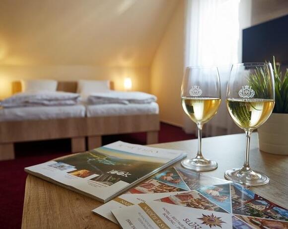 Gemütliches Dachzimmer mit Doppelbett, Schrägdach und warmen Lampen; Tisch mit zwei Weißweingläsern, Broschüren und Pflanze.