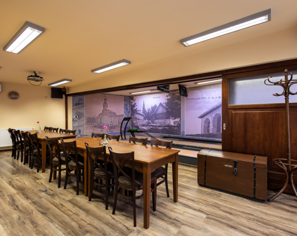Pohodlná hotelová soukromá jídelna a zasedací místnost s dlouhým dřevěným stolem, židlemi, stropním projektorem a historickou fotostěnou