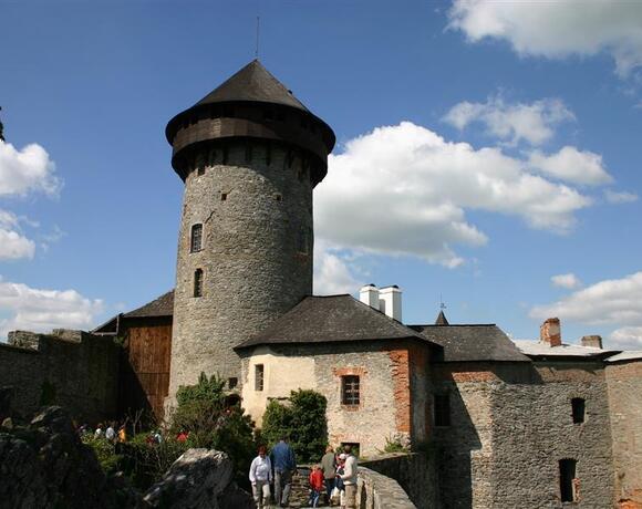 Středověký kamenný hrad s kulatou věží a nádvořím; návštěvníci procházejí po kamenné cestě pod modrou oblohou