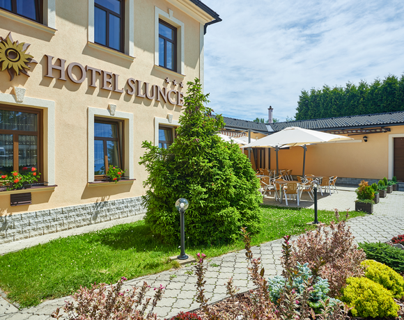 Pfirsichfarbene Hotelfassade mit Sonnenlogo, Blumenkästen, gepflastertem Innenhof und schattiger Außenterrasse.