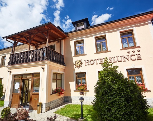 Slunná fasáda butikového hotelu s dřevěným balkonem, květinovými truhlíky u oken, skleněným vchodem a třemi hvězdami