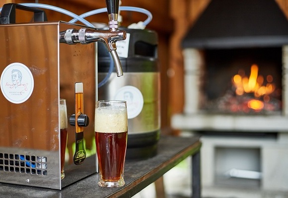 Jantarové řemeslné pivo na čepu v hotelovém baru, pěnivá sklenice vedle soudku a hřejivého krbu