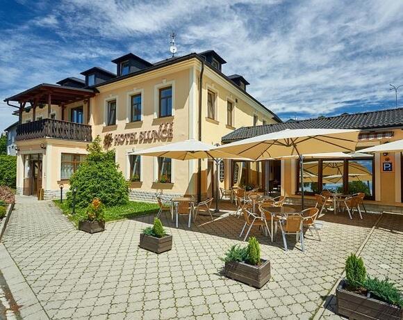 Dvoupodlažní žlutý hotel s dřevěným balkonem a slunnou dlážděnou terasou s stolky, slunečníky a květinami v květináčích.