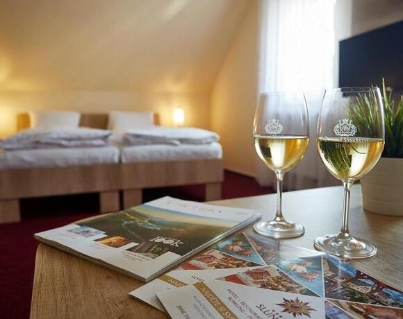 Útulný podkrovní pokoj se dvěma lůžky a tlumenými nočními lampami; na stole dvě sklenky bílého vína, brožury a květina v květináči