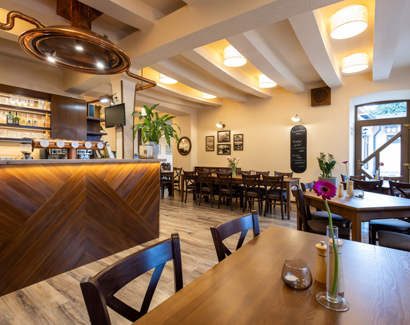 Příjemná hotelová jídelna s dřevěným barem, měděným krytem nad barem, dlouhými společnými stoly, odkrytými trámy a růžovým květem na stole.