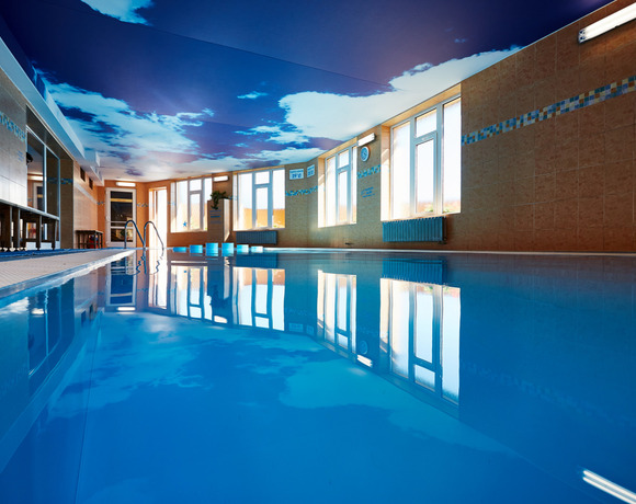 Vnitřní hotelový bazén s nebeskou malbou na stropě, velkými okny, dlaždicovým okrajem a zrcadlící klidnou hladinou.