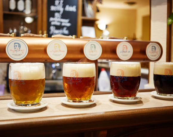 Čtyři řemeslná piva s bohatou pěnou na dřevěném baru před měděným výčepem s logy, teplá útulná hospodská atmosféra