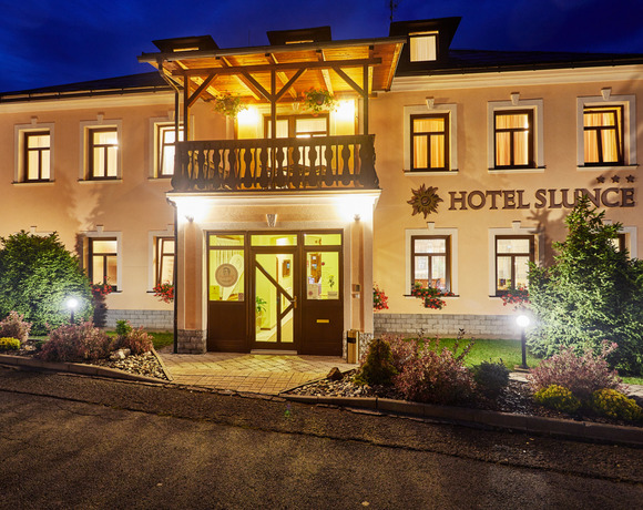 Večerní osvětlená dvoupodlažní fasáda hotelu s dřevěným balkonem, vstupem, květinami ve truhlících a upravenou zahrádkou