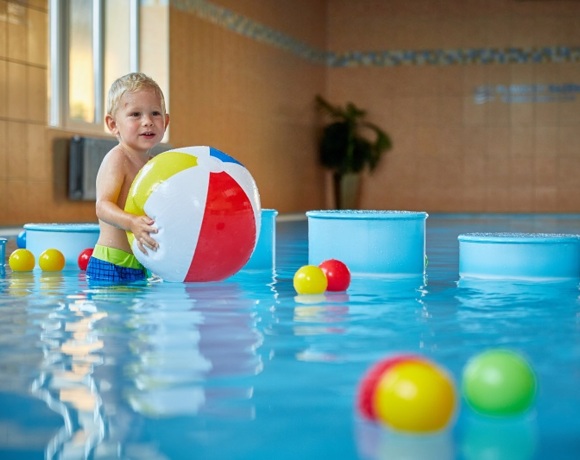 Batole si hraje s barevným míčem v mělkém dětském bazénu s plovoucími hračkami ve Wellness & Aquacentru Slunce v Hotelu Slunce v Rýmařově v Jeseníkách. Vnitřní bazén a dětská herní zóna jsou ideální pro rodiny s dětmi během pobytu v hotelu nebo wellness návštěvy.