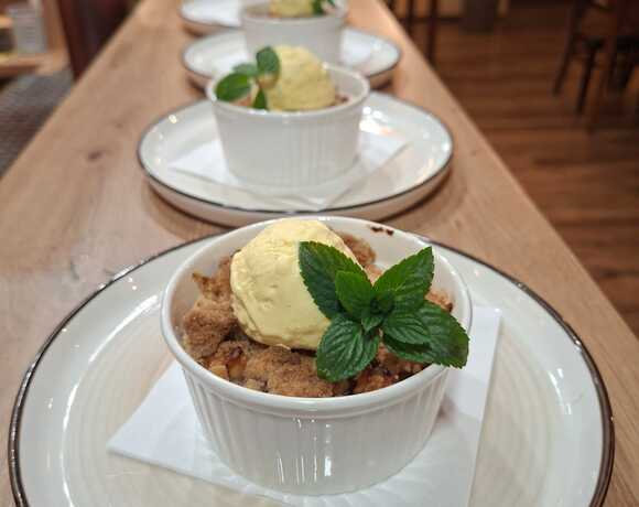 Ovocný crumble podávaný v bílém ramekinu s vanilkovou zmrzlinou a čerstvou mátou na talíři u dřevěného baru v restauraci Hotelu Slunce v Rýmařově v Jeseníkách. Domácí dezert z hotelové restaurace, ideální sladká tečka po dobrém jídle a pivu z pivovaru Morous.