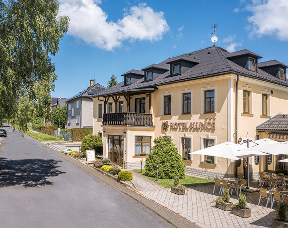 Budova Hotelu Slunce Rýmařov v Jeseníkách s balkonem, venkovní terasou se slunečníky a klidnou polohou v ulici lemované stromy.