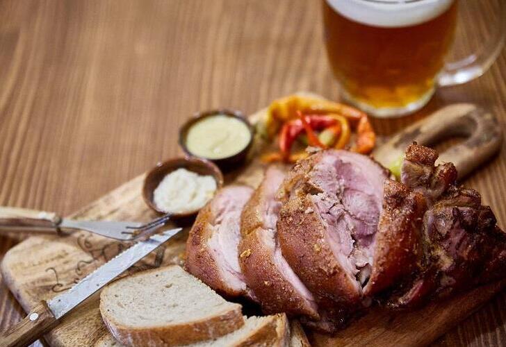 Scheiben von gebratenem Schweinshaxenfleisch, serviert auf einem Holzbrett mit frischem Brot, Senf, Meerrettich und eingelegter Paprika, dazu ein halber Liter frisch gezapftes Bier aus der Familienbrauerei Morous. Eine traditionelle tschechische Spezialität, serviert im Restaurant des Hotels Slunce in Rýmařov im Altvatergebirge (Jeseníky).