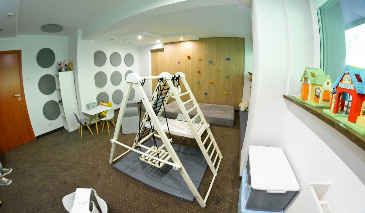 Sala zabaw hotelowa z drewnianą drabinką i huśtawką, miękkimi matami, ścianką wspinaczkową, stolikiem i zabawkami.