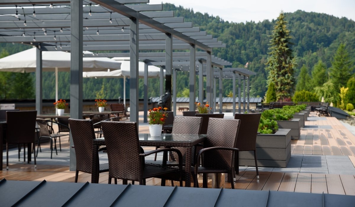 Taras restauracyjny z rattanowymi stolikami, pergolą ze światłami, parasolami i donicami z kwiatami; widok na zalesione góry.