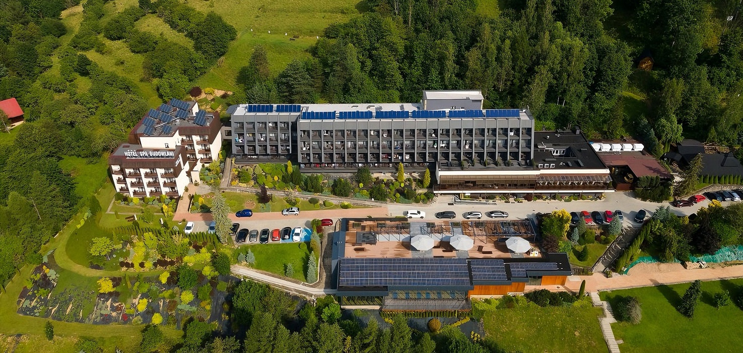Widok z lotu ptaka na górski hotel z balkonami, tarasem spa z parasolami, panelami słonecznymi i parkingiem wśród lasu.