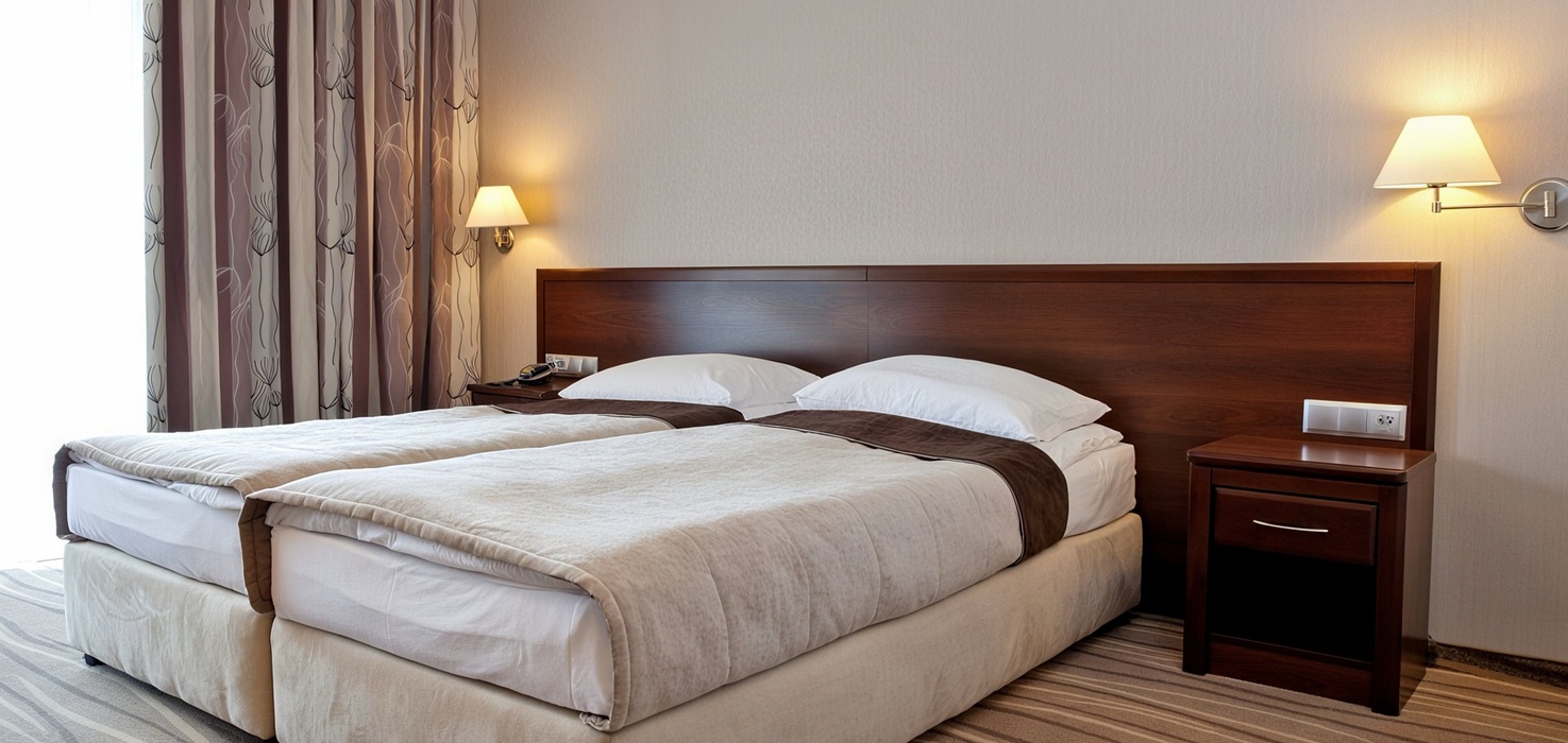 Dwuosobny pokój z dwoma łóżkami, drewnianym zagłówkiem, miękkimi narzutami, nocnymi stolikami, lampami i wzorzystymi zasłonami