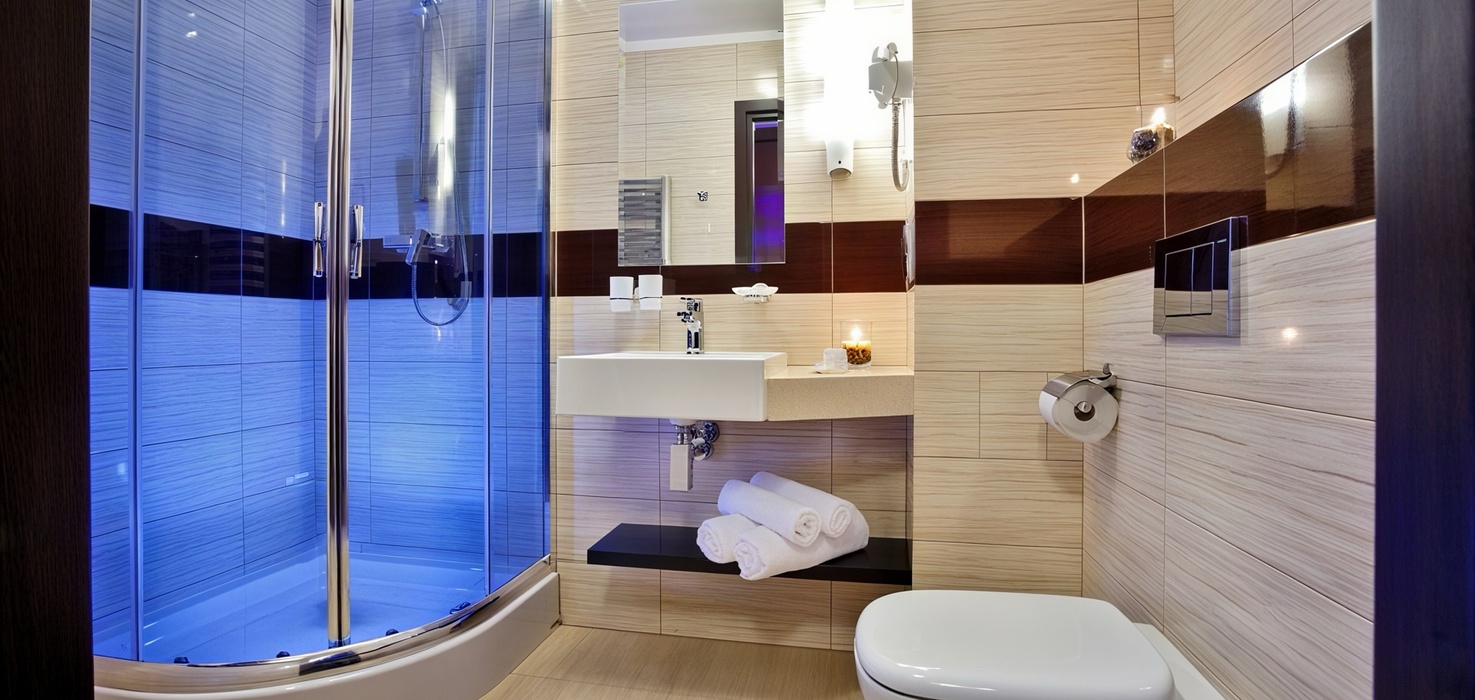 Hotelowa łazienka z niebiesko podświetloną kabiną prysznicową, podwieszaną umywalką, ręcznikami i ścienną toaletą.