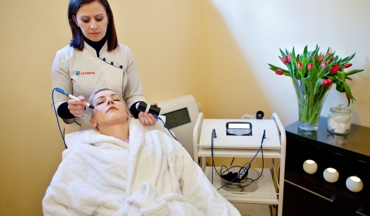 Kosmetyczka wykonuje zabieg na twarz urządzeniem, klientka w białym szlafroku na leżance, wazon z czerwonymi tulipanami.