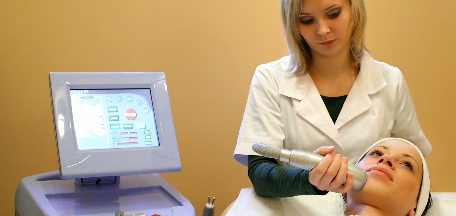 Profesjonalny zabieg twarzy: kosmetyczka używa urządzenia na twarz klientki leżącej z opaską, obok widać monitor sprzętu.