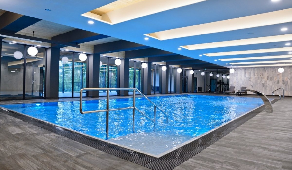Nowoczesny kryty basen hotelowy z niebieską wodą, stalowymi poręczami, przeszklonymi ścianami, okrągłymi lampami i leżakami