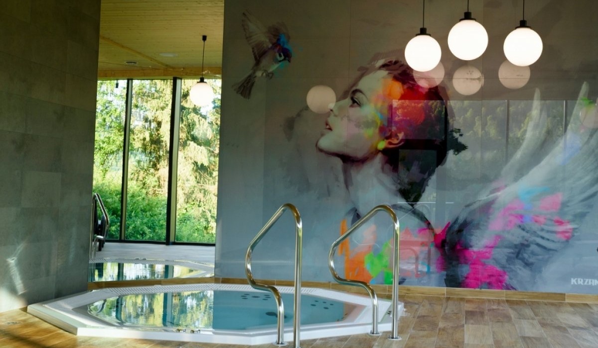 Wnętrze spa: jacuzzi z metalowymi poręczami, mural kobiety z kolorowymi skrzydłami, duże okna na zieleń.