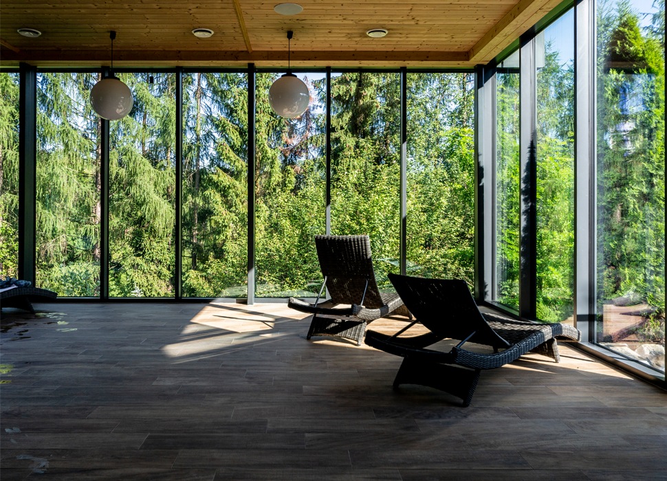 Pokój relaksu spa z przeszklonymi ścianami, wiklinowymi leżakami, drewnianym sufitem i panoramicznym widokiem na las.