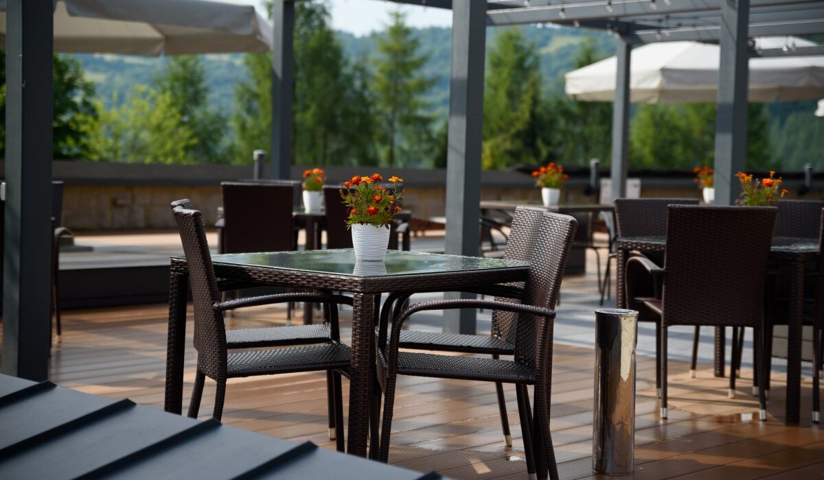 Taras hotelowej restauracji z rattanowymi stolikami, doniczkami z kwiatami, pergolą, parasolami i widokiem na leśne wzgórza.