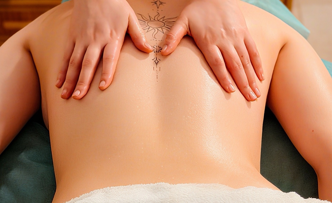 Relaksacyjny masaż pleców z olejkiem w spa, terapeutka masuje okolice łopatek nad tatuażem słońca, miękki biały ręcznik.