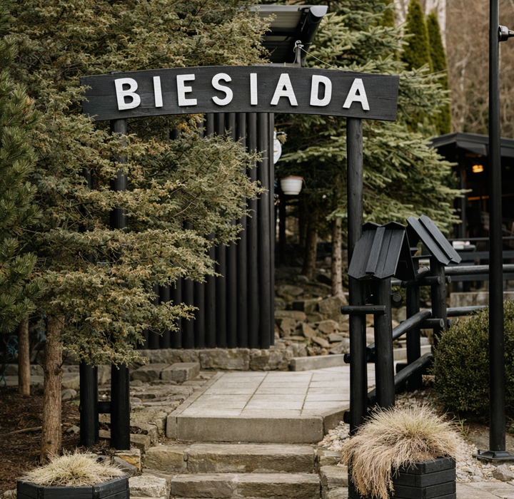Wejście do rustykalnej restauracji Biesiada: drewniana brama, kamienne schody, iglaste drzewa i donice