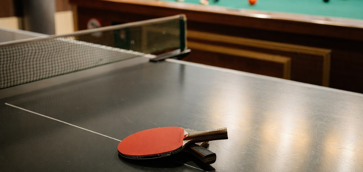 Stół do ping-ponga z dwiema rakietkami na środku, w tle stół bilardowy — przytulna sala gier hotelowych