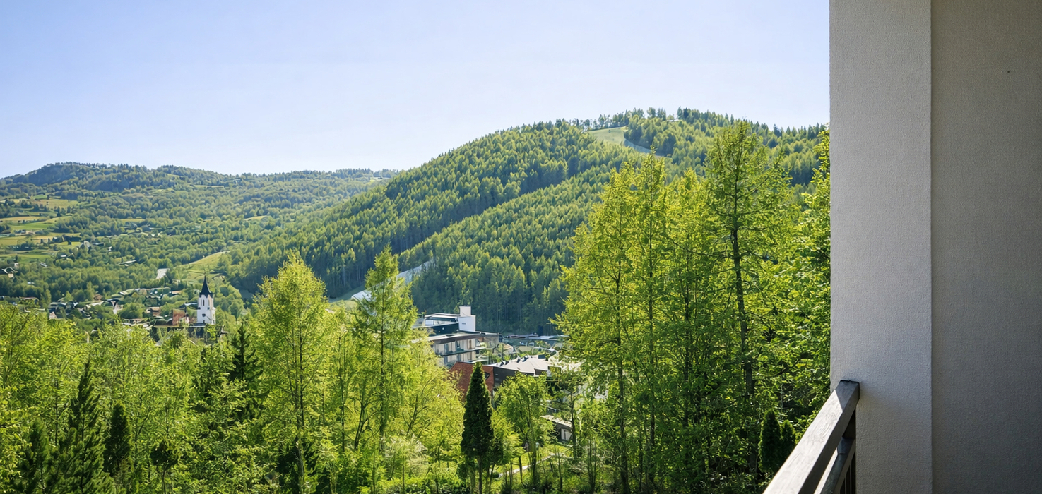 Widok z hotelowego balkonu na zielone wzgórza, gęsty las i malowniczą wioskę z wieżą kościelną.