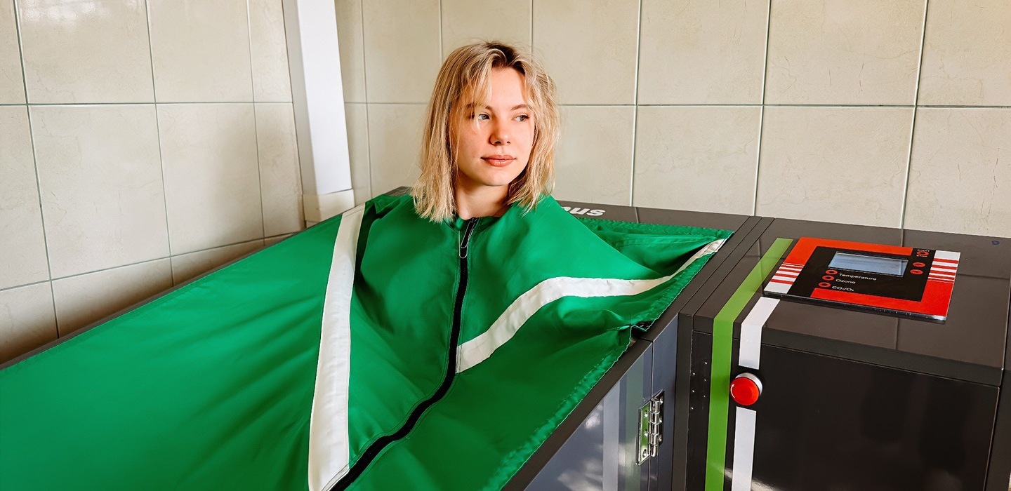 Kobieta relaksująca się w zielonej kapsule zabiegowej do odnowy z panelem sterowania, w jasnym, kafelkowanym pokoju spa