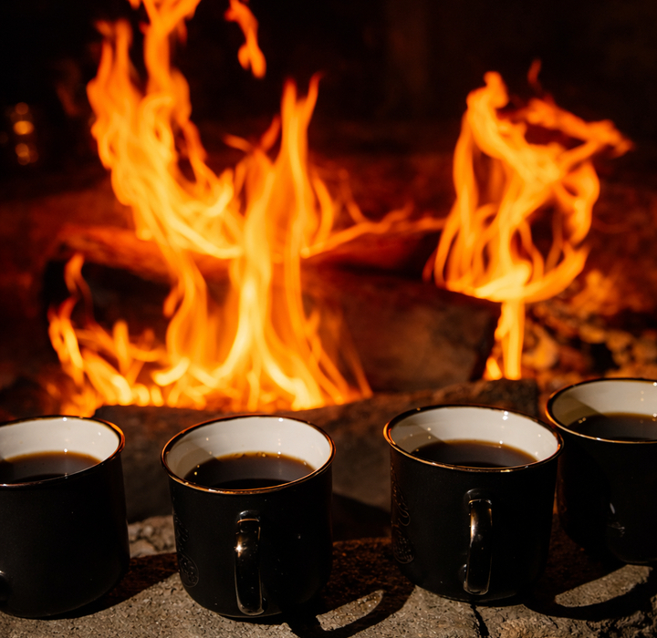 Cztery czarne kubki z gorącym napojem ustawione na kamieniu przed trzaskającym kominkiem, przytulna atmosfera