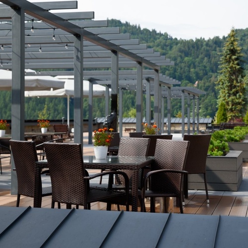 Taras restauracyjny z rattanowymi stolikami, pergolą ze światłami, parasolami i donicami z kwiatami; widok na zalesione góry.