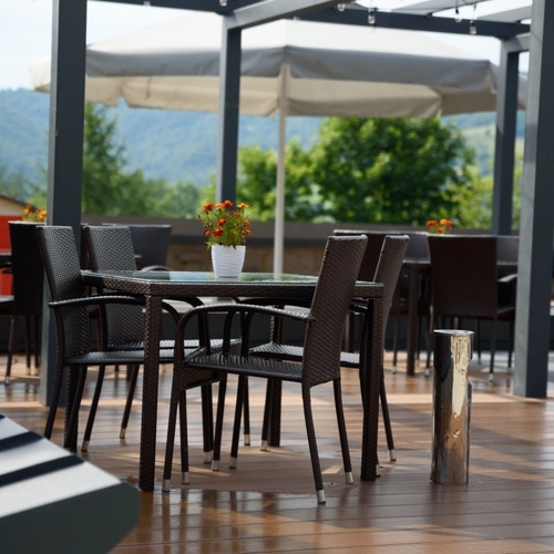 Taras restauracyjny z wiklinowymi krzesłami i stolikami, kwiaty na szklanym blacie, parasol i górski widok.