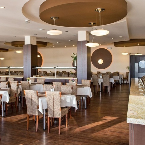 Przestronna restauracja hotelowa z białymi obrusami, ustawionymi stolikami, bufetem po prawej i ciepłym oświetleniem