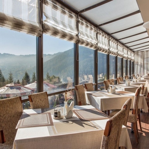 Restauracja z przeszkloną werandą i ustawionymi stolikami ze śniadaniowymi nakryciami, panoramiczny widok na góry.