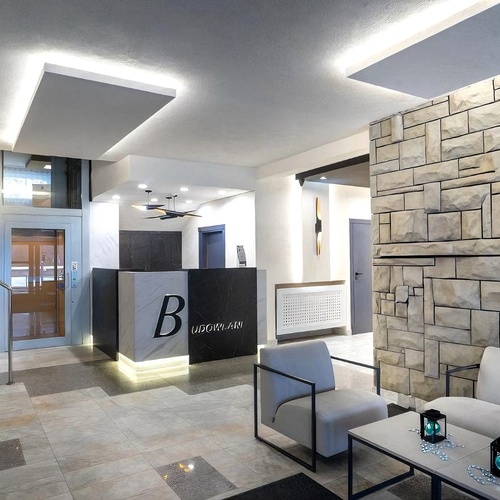 Eleganckie lobby z marmurowymi schodami i windą, podświetloną recepcją z logo B, kamienną ścianą i strefą wypoczynku.