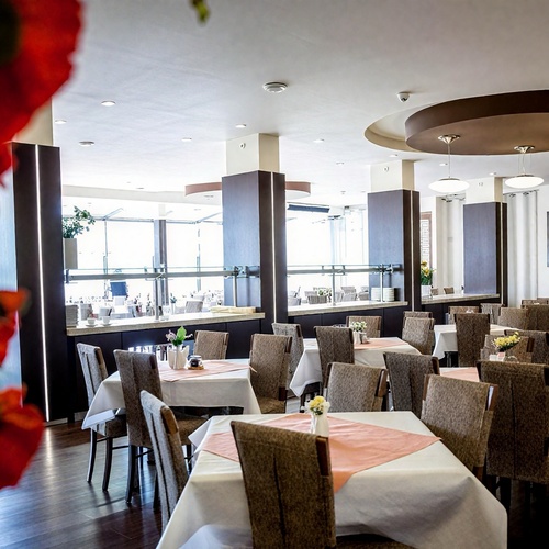 Przestronna restauracja hotelowa z białymi obrusami, łososiowymi serwetkami, stolikami, bufetem przy oknach i kwiatami.