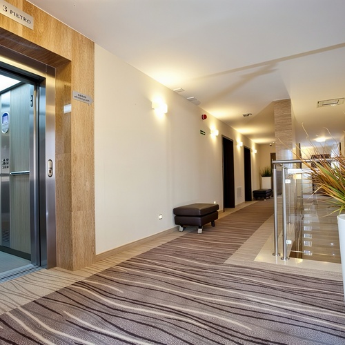 Korytarz hotelowy z otwartą windą, paskowanym wykładzinem, skórzanymi ławeczkami i dużą donicą przy szklanej balustradzie.