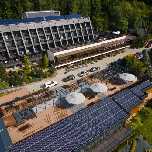 Hotel z tarasem wypoczynkowym z parasolami, dużymi panelami słonecznymi, balkonami pokojowymi i leśnym otoczeniem.