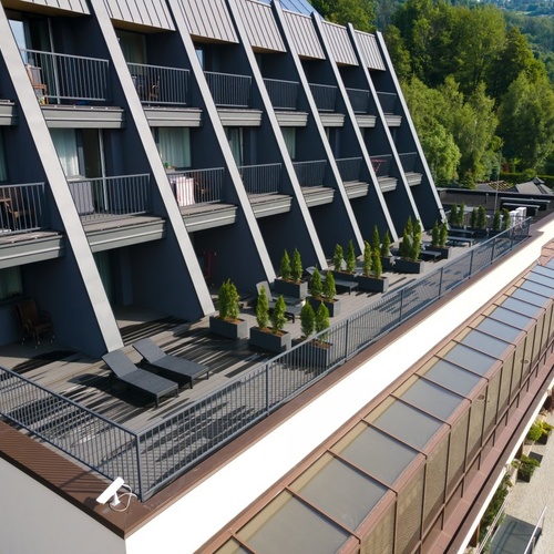 Nowoczesny hotel z pochylonymi balkonami, tarasem z leżakami i donicami, widok na las.