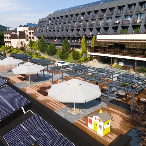 Taras hotelowy z pergolami, parasolami, leżakami i małym domkiem zabaw; nowoczesny budynek z balkonami i panelami słonecznymi