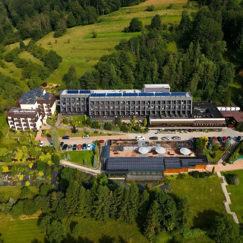 Widok z lotu ptaka na górski hotel z balkonami, tarasem spa z parasolami, panelami słonecznymi i parkingiem wśród lasu.