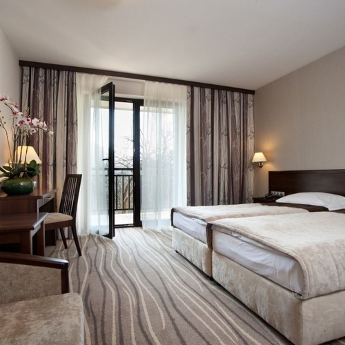 Pokój z dwoma pojedynczymi łóżkami, balkonem przeszklonym, biurkiem z orchideą, ściennym telewizorem i lampami nocnymi.