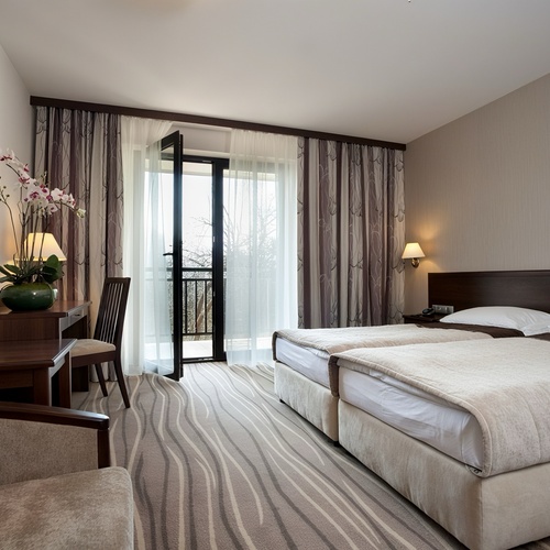 Elegancki pokój z dwoma pojedynczymi łóżkami, balkonem przez przeszklone drzwi, biurkiem z orchideą, telewizorem i lampami nocnymi.